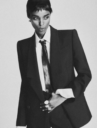 Monochrome portrait of a model in a tailored black suit with white shirt and dark tie against a seamless white background.