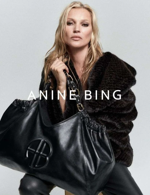 Kate Moss poses with a large black leather handbag, modelling a dark fur jacket and black leather pants against a neutral backdrop.
