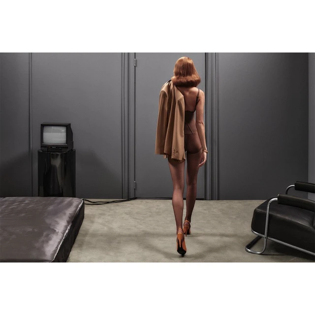 A woman in lingerie and heels walks in an interior space with a jacket over her shoulder. Retro television and chair.