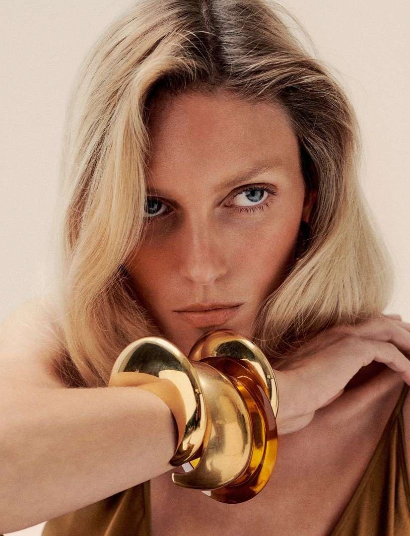 Close-up portrait of a fair-skinned woman with blonde wavy hair and gold bangle bracelets, against a neutral-toned background.