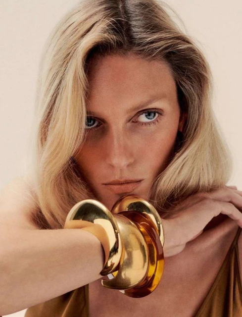 Close-up portrait of a fair-skinned woman with blonde wavy hair and gold bangle bracelets, against a neutral-toned background.