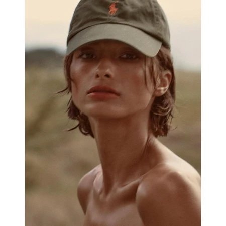 Close-up portrait of a woman wearing an olive green baseball cap outdoors against a soft natural background.