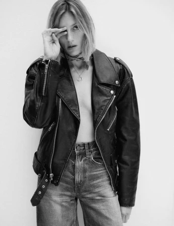 Model in black leather jacket and jeans with hand near face against white background