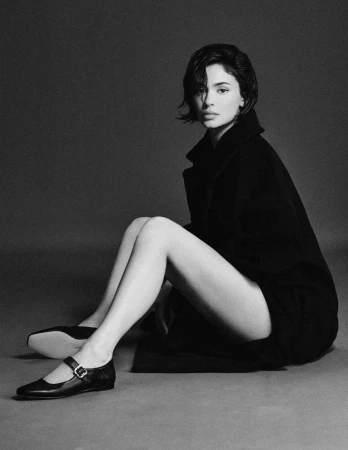 Monochrome portrait of a woman with short dark hair wearing a long black coat and flat shoes, seated on a grey studio floor.