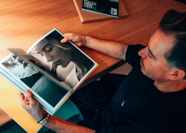 Man reads black and white magazine on wooden table, casual wear, natural light, modern interior, calm mood, showcasing design and lifestyle.