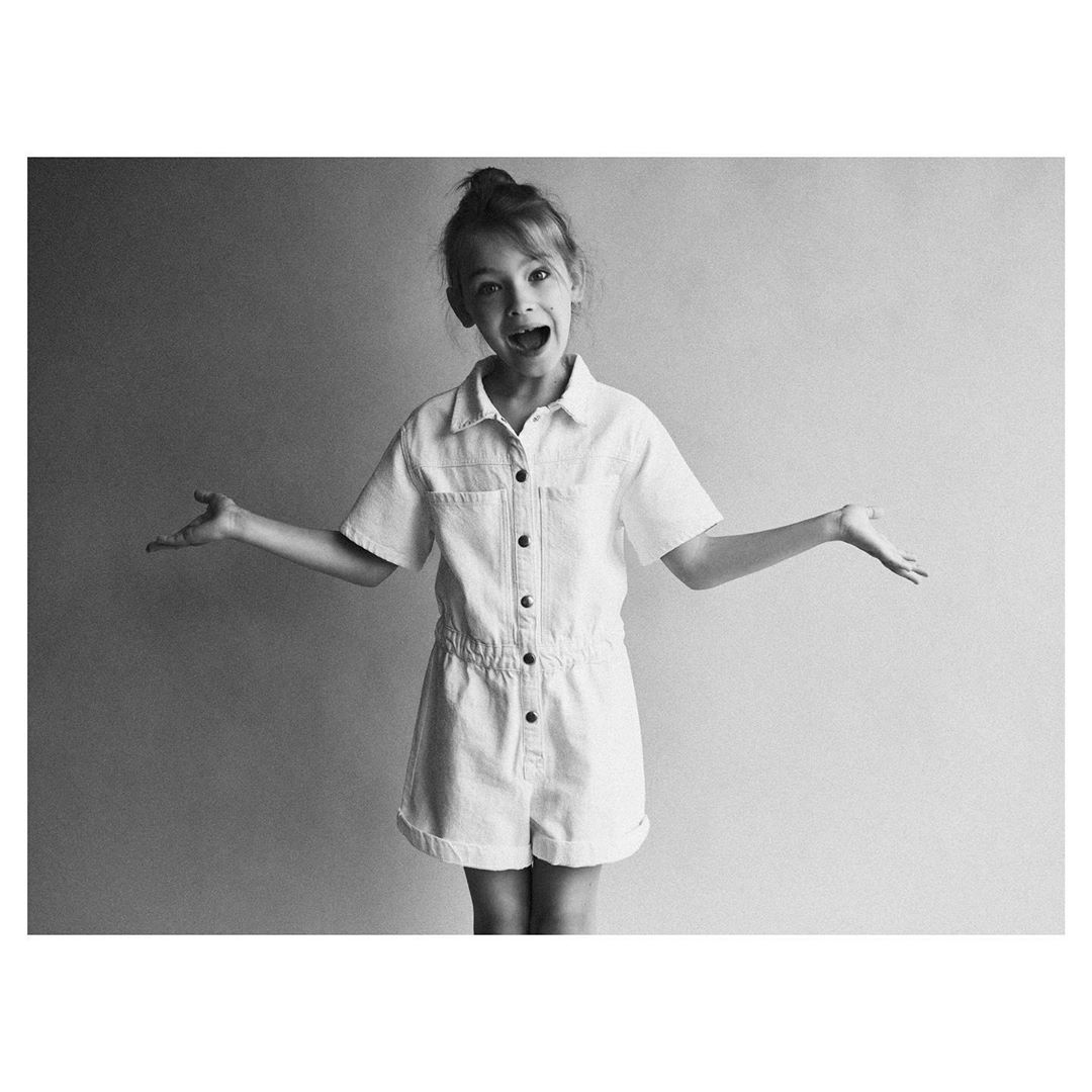 A young child with a topknot and wearing a white romper poses with an open mouth and outstretched arms against a neutral backdrop.