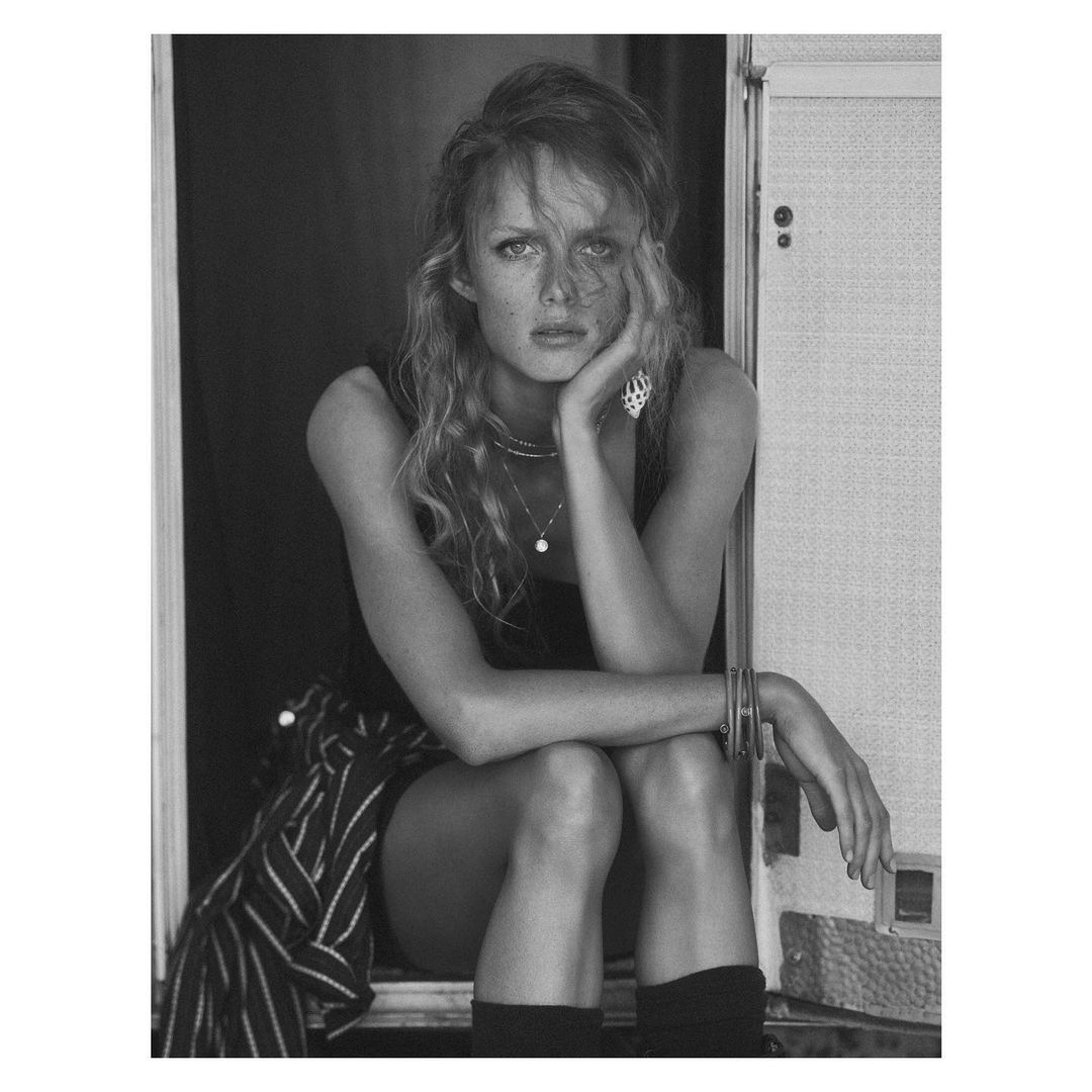 Monochrome portrait of a young woman with long curly hair sitting in front of an open doorway with striped fabric and jewelry.