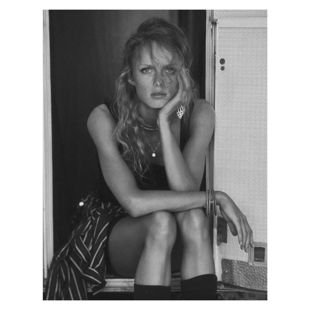 Monochrome portrait of a young woman with long curly hair sitting in front of an open doorway with striped fabric and jewelry.