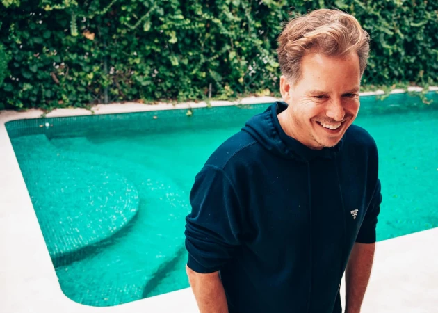 Man wearing a dark blue hoodie smiling near an outdoor swimming pool with lush green foliage in the background