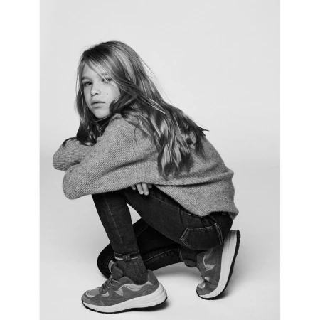 Young female model crouching in a grey ribbed sweater, dark jeans, and sneakers against a plain background