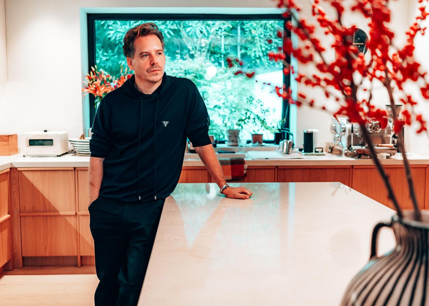 Adult male in dark hoodie and pants leans on white countertop in a modern kitchen. Natural light from a window illuminates the scene.