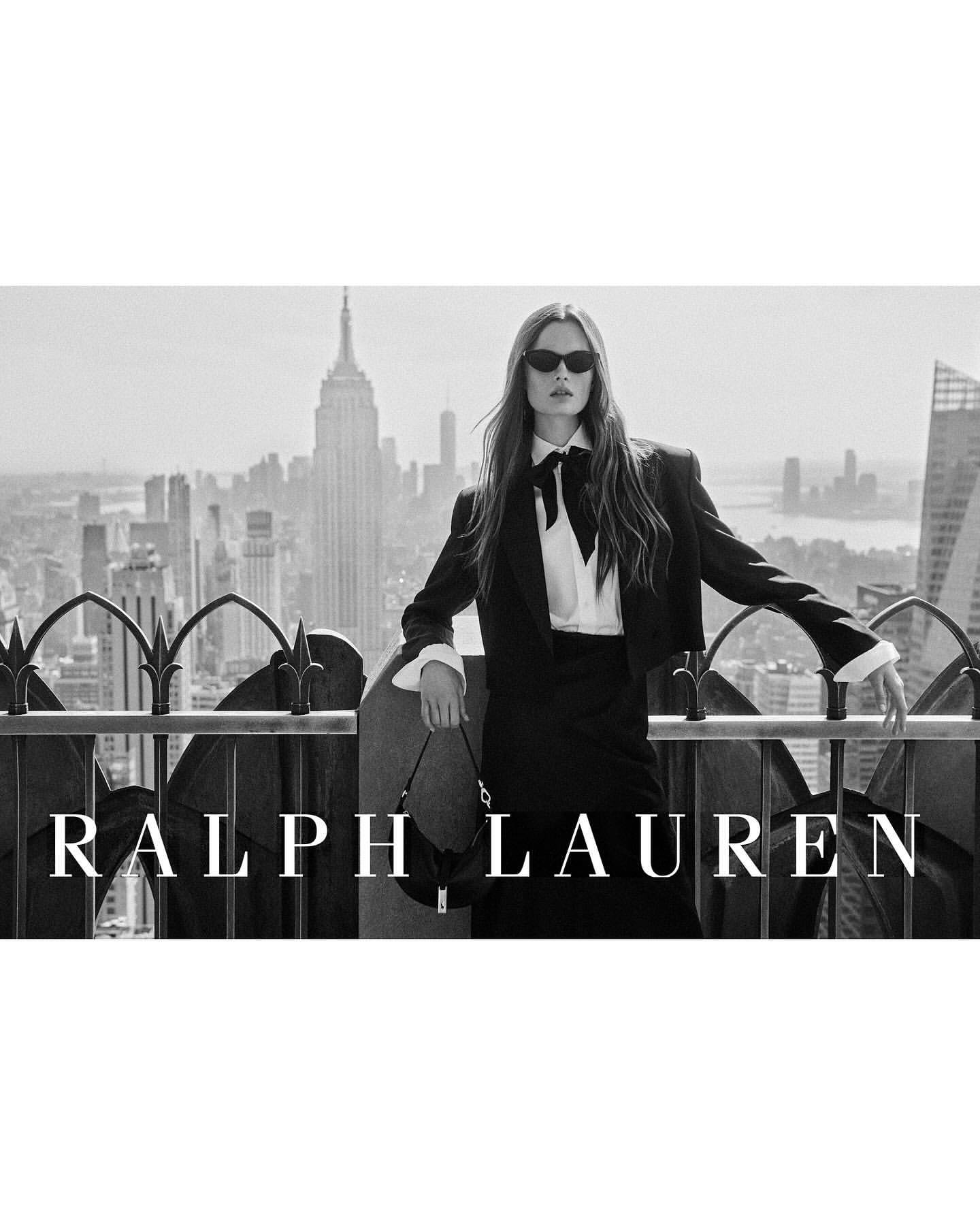 Model in black suit jacket and white collared shirt with a black bow holding a handbag on a rooftop overlooking a city skyline