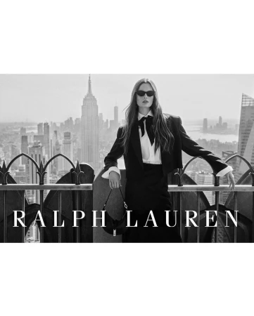 Model in black suit jacket and white collared shirt with a black bow holding a handbag on a rooftop overlooking a city skyline