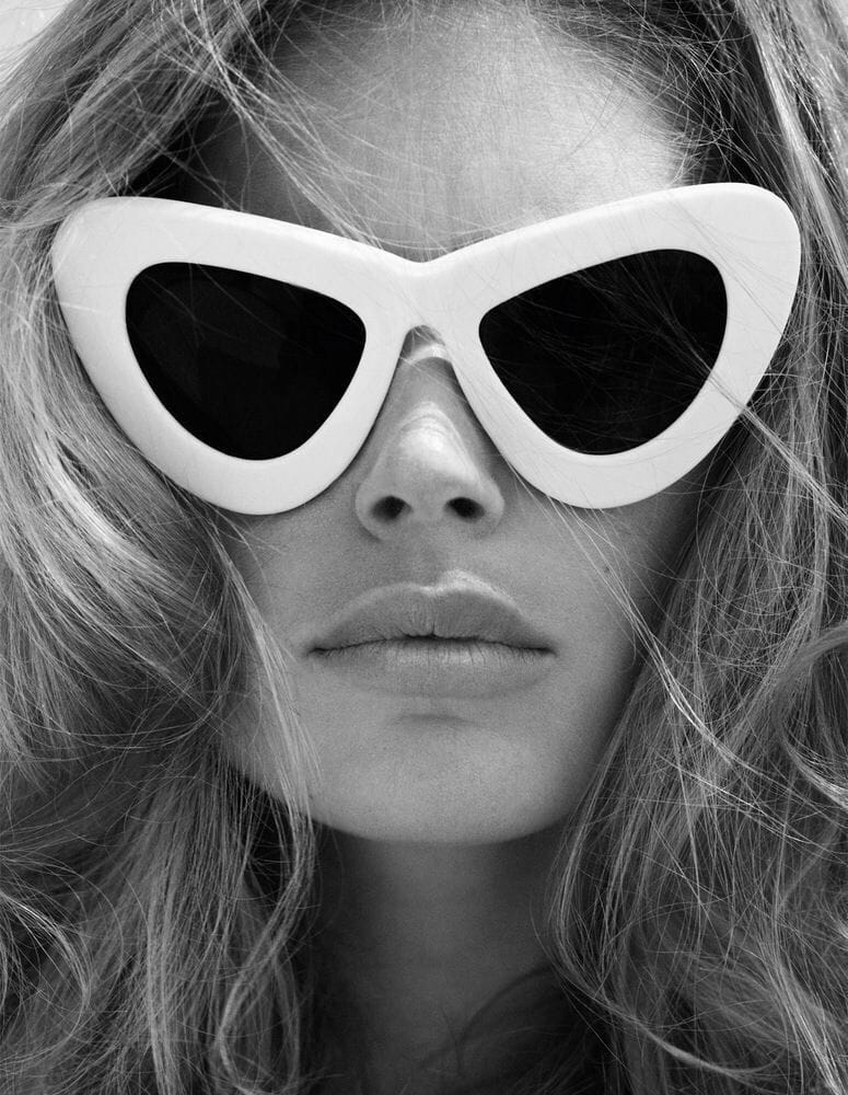 Close-up portrait of a woman wearing oversized white cat-eye sunglasses, showcasing a high-fashion, minimalist aesthetic in black and white.