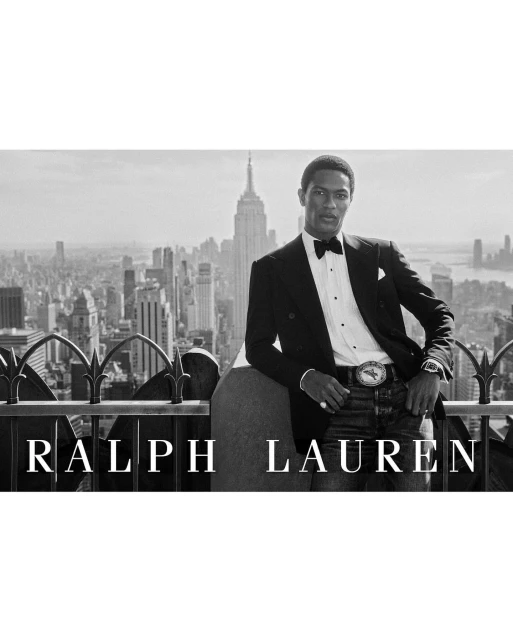 Black and white portrait of a black male model in a tuxedo, posing outdoors with a cityscape and the Empire State Building.