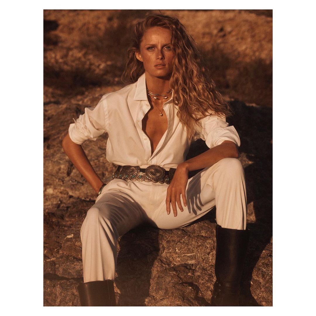 A woman with wavy blonde hair sits on rocks outdoors, wearing a white shirt, pants, silver belt, and black boots in a fashion style.