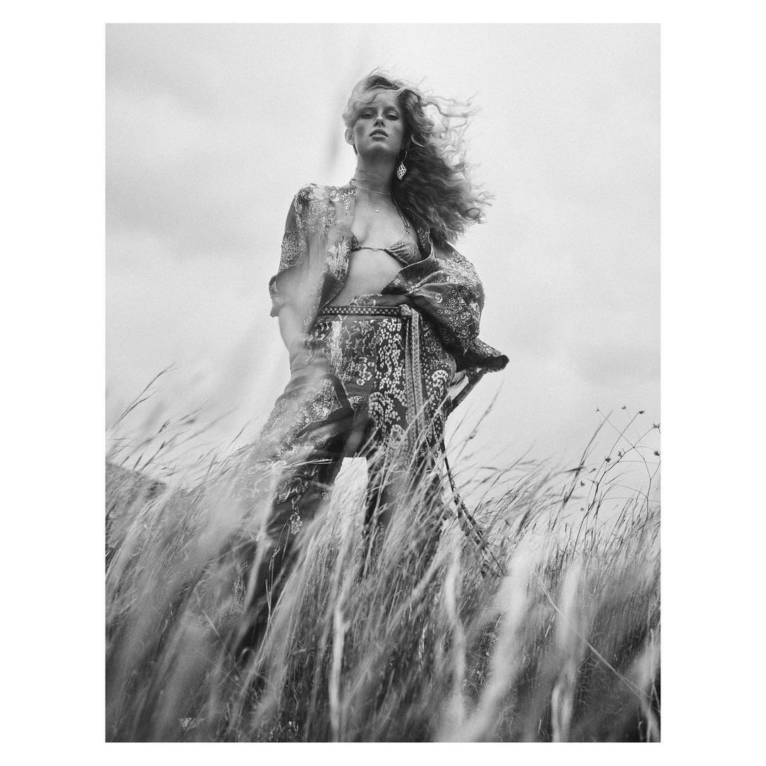 Black and white portrait of woman in patterned outfit posing in a field of tall grass under an overcast sky in a fashion editorial.