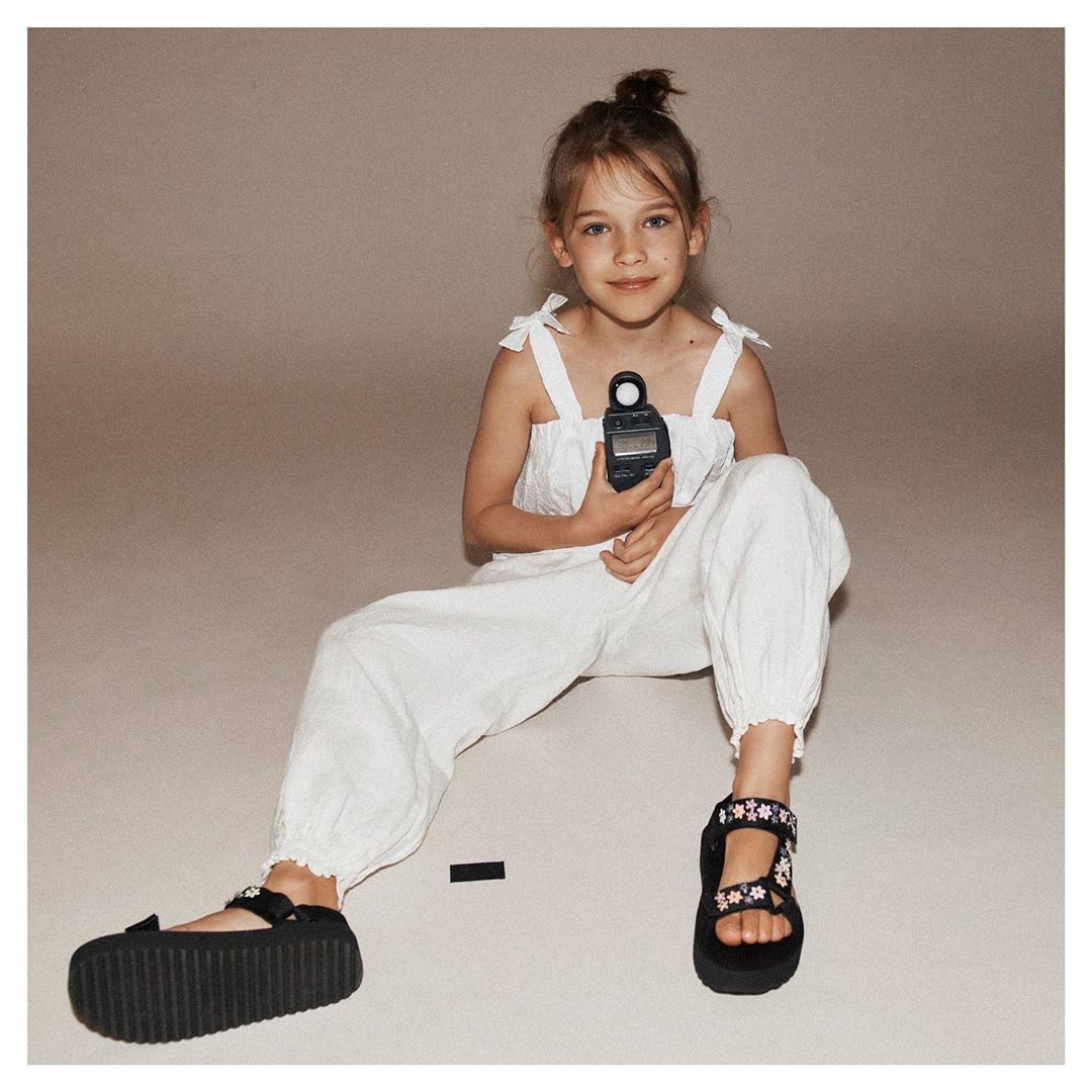 A young girl in a white outfit sits cross-legged indoors holding a light meter, wearing sandals with flower details on a neutral backdrop.