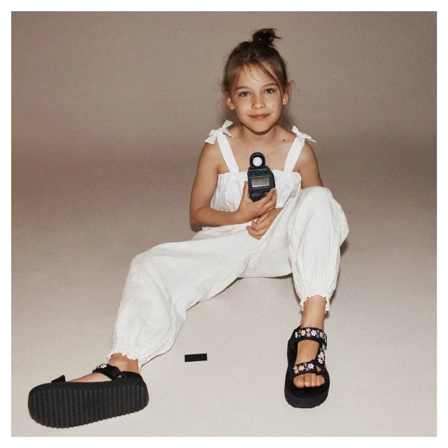 A young girl in a white outfit sits cross-legged indoors holding a light meter, wearing sandals with flower details on a neutral backdrop.