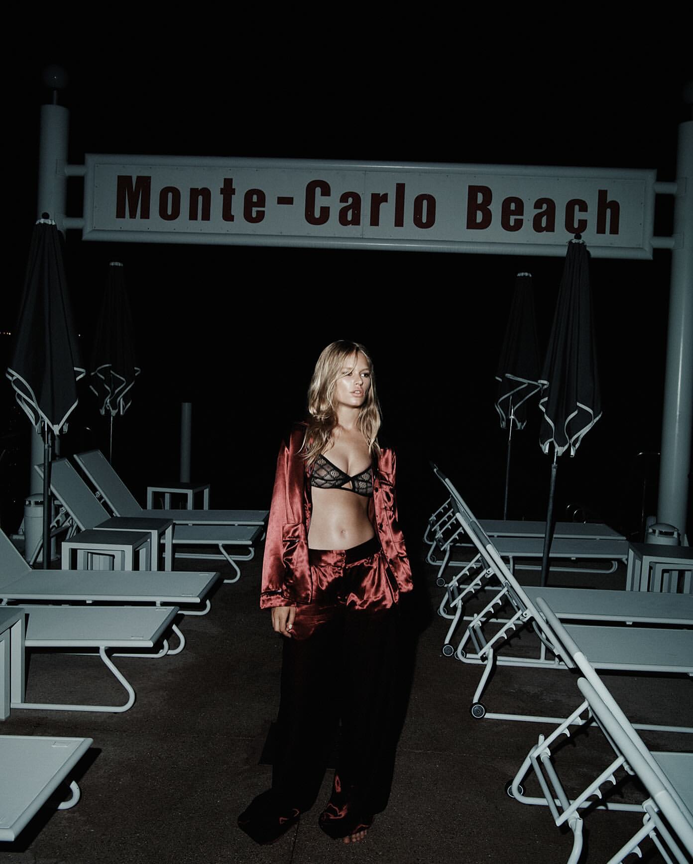 A blonde woman stands on a beach at night, posing in lingerie and a silk robe, with lounge chairs under a sign reading Monte-Carlo Beach.