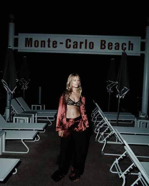 A blonde woman stands on a beach at night, posing in lingerie and a silk robe, with lounge chairs under a sign reading Monte-Carlo Beach.