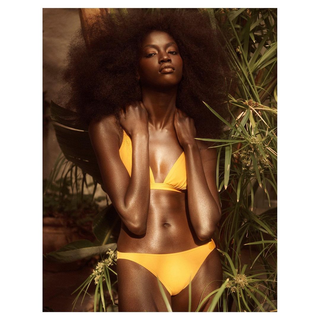 A dark-skinned woman with natural hair poses in a yellow bikini top and bottom, standing among green plants, with soft, warm lighting.