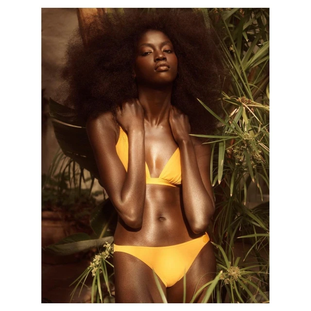 A dark-skinned woman with natural hair poses in a yellow bikini top and bottom, standing among green plants, with soft, warm lighting.