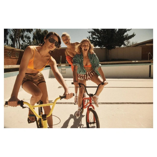 Three young women ride BMX bicycles in an empty swimming pool on a sunny day. The women are wearing summery clothing and smiling.