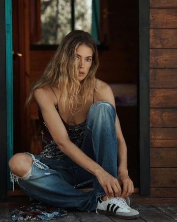 Blonde woman in a black top and ripped jeans sitting and tying a black and white Adidas sneaker in front of a cabin.