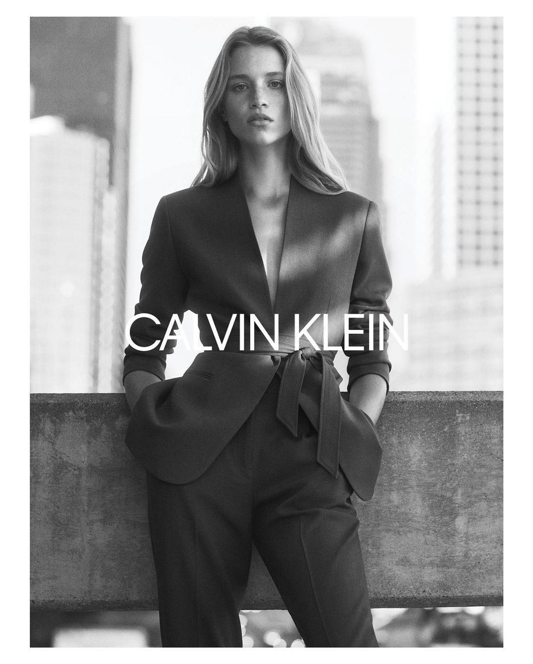 Black and white portrait of a female model wearing a tailored belted blazer and trousers against an urban rooftop background.