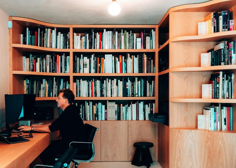 Adult male works at a computer on a desk surrounded by a tall bookcase filled with books in a warm, well-lit, indoor environment.