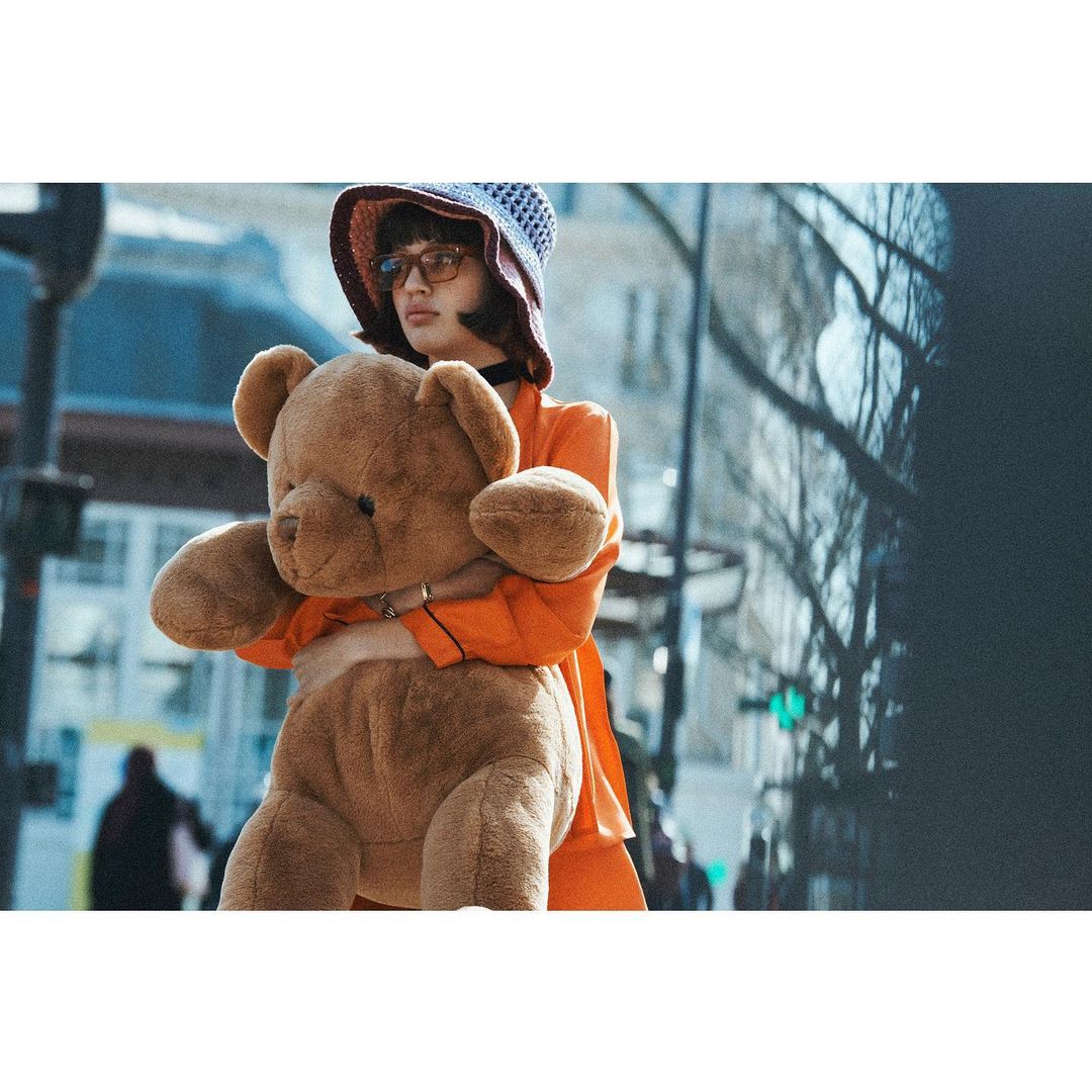 A young adult woman in an orange jacket, wearing a hat and glasses, holds a large brown teddy bear in an urban street setting.