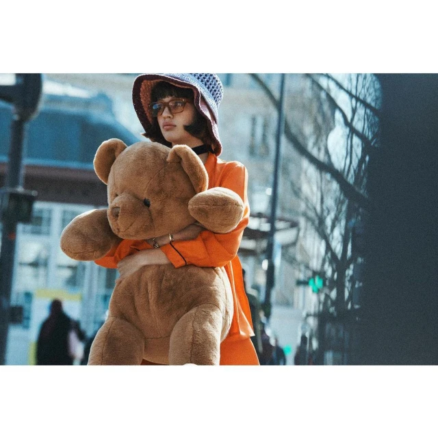 A young adult woman in an orange jacket, wearing a hat and glasses, holds a large brown teddy bear in an urban street setting.