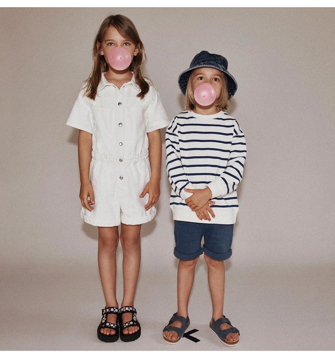 Two kids blowing bubblegum bubbles. A girl in a white romper and a boy in a striped shirt and blue bucket hat stand in studio.