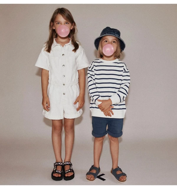 Two kids blowing bubblegum bubbles. A girl in a white romper and a boy in a striped shirt and blue bucket hat stand in studio.