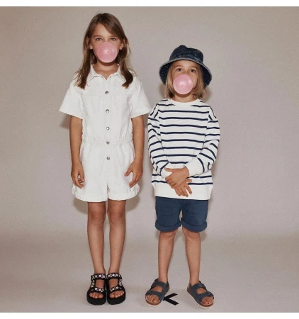 Two kids blowing bubblegum bubbles. A girl in a white romper and a boy in a striped shirt and blue bucket hat stand in studio.