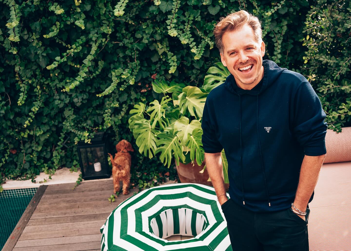 Cheerful adult man in casual wear standing outdoors on a deck with a dog, lush green plants, and a pool in the background.