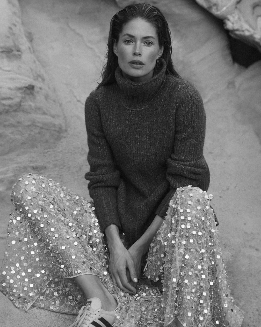 Woman in a turtleneck sweater and sequined skirt sits on a sandy rock formation, posing in a black-and-white fashion portrait.