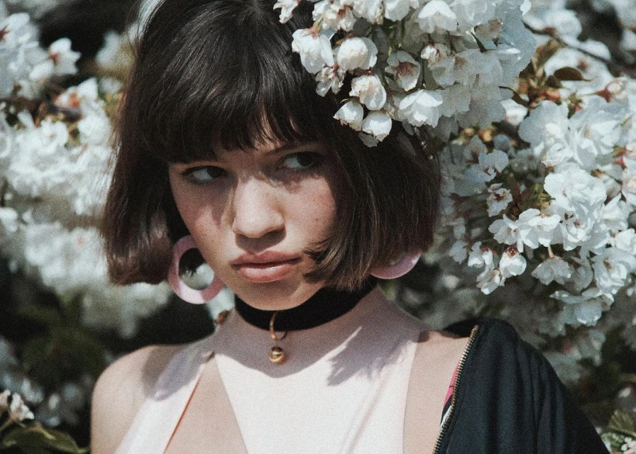 Close-up portrait of a young woman with dark hair wearing hoop earrings and a black choker, standing in front of a flowering tree.