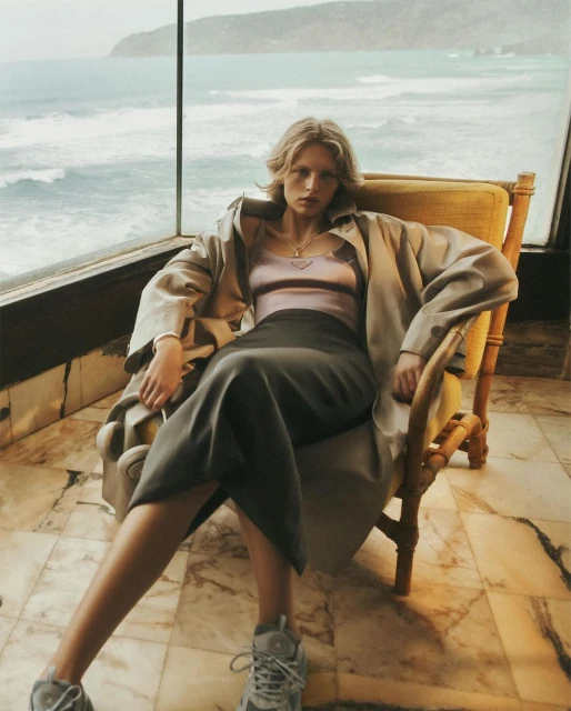 A relaxed adult woman sits in a yellow armchair wearing a trench coat, top, skirt, and sneakers with an ocean view in the background.