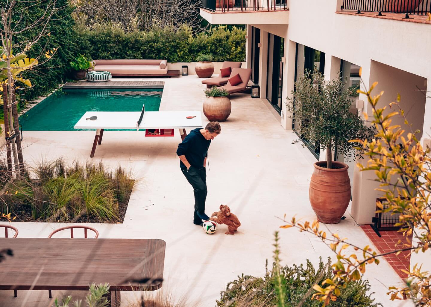A man plays soccer with a dog on a patio near a pool. The scene features outdoor furniture and lush greenery in the background.