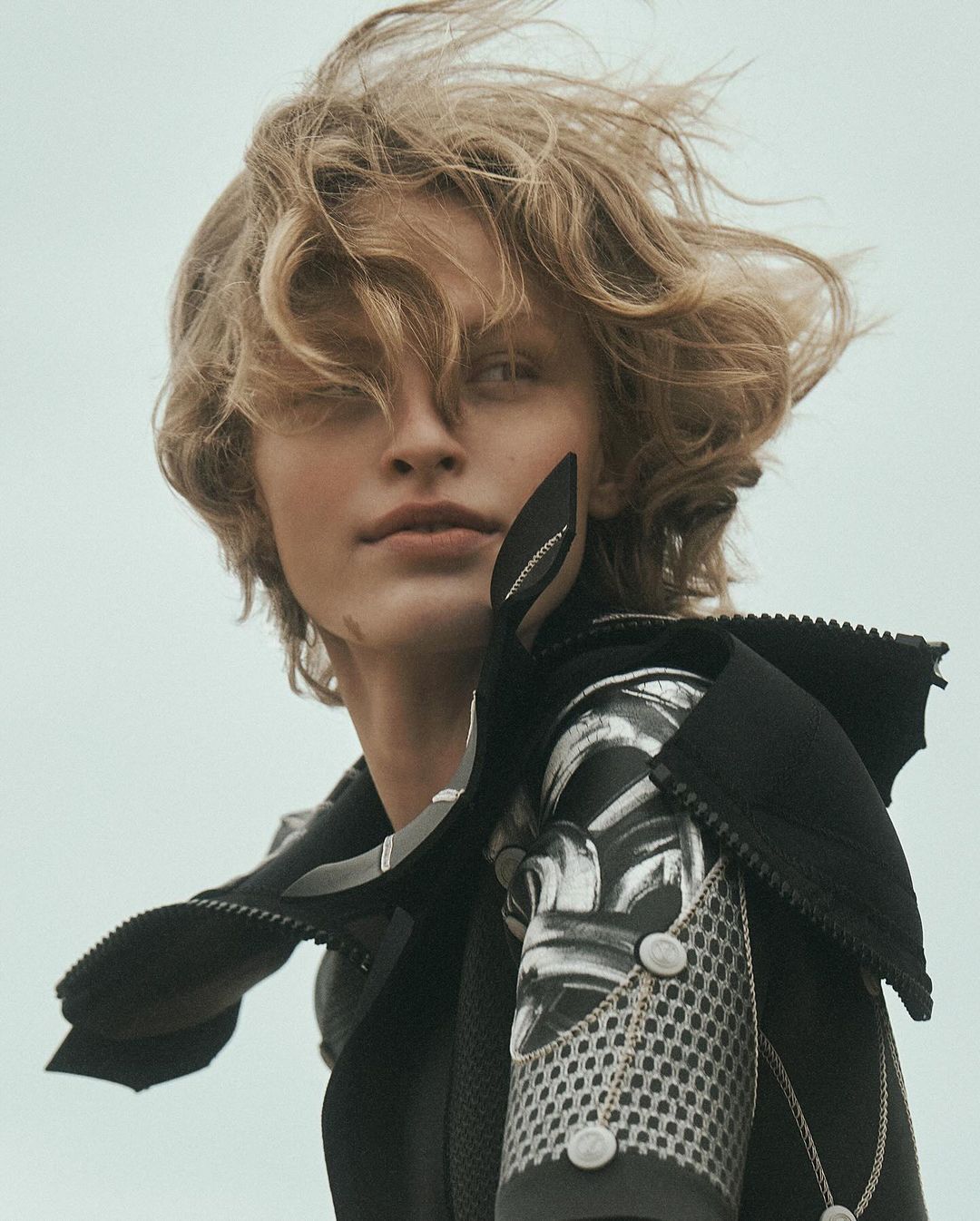 Close-up fashion shot of a stylish adult with wavy blonde hair wearing a black jacket against a bright neutral background.