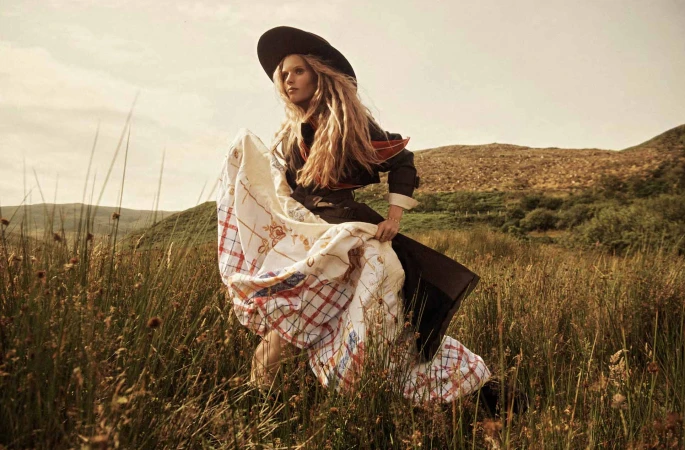 Model in wide-brimmed hat holding patterned blanket in grassy field by Lachlan Bailey