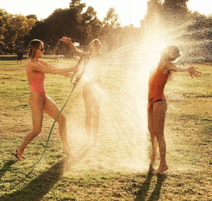 Three women in swimsuits outdoors on grass with trees; one woman holding hose spraying water onto another, bright sunny day.
