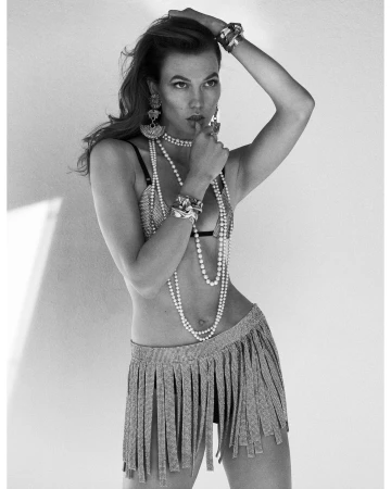 Black and white portrait of a woman wearing layered pearl necklaces, bracelets, and a fringe skirt against a plain background.