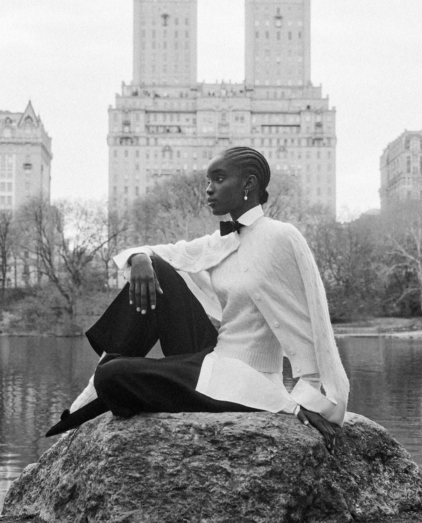 Elegant woman poses on a rock in a park with city buildings in the background. She wears a light sweater, black pants, and a bow tie.