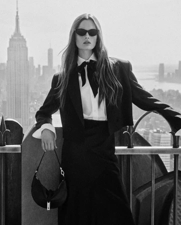 Model in black suit and sunglasses holding handbag on rooftop with city skyline backdrop by Lachlan Bailey