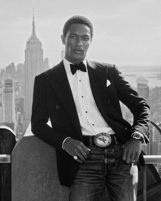 A black-and-white portrait of an African-American male model in a tuxedo with an ornate belt buckle, posing in front of the New York City skyline.