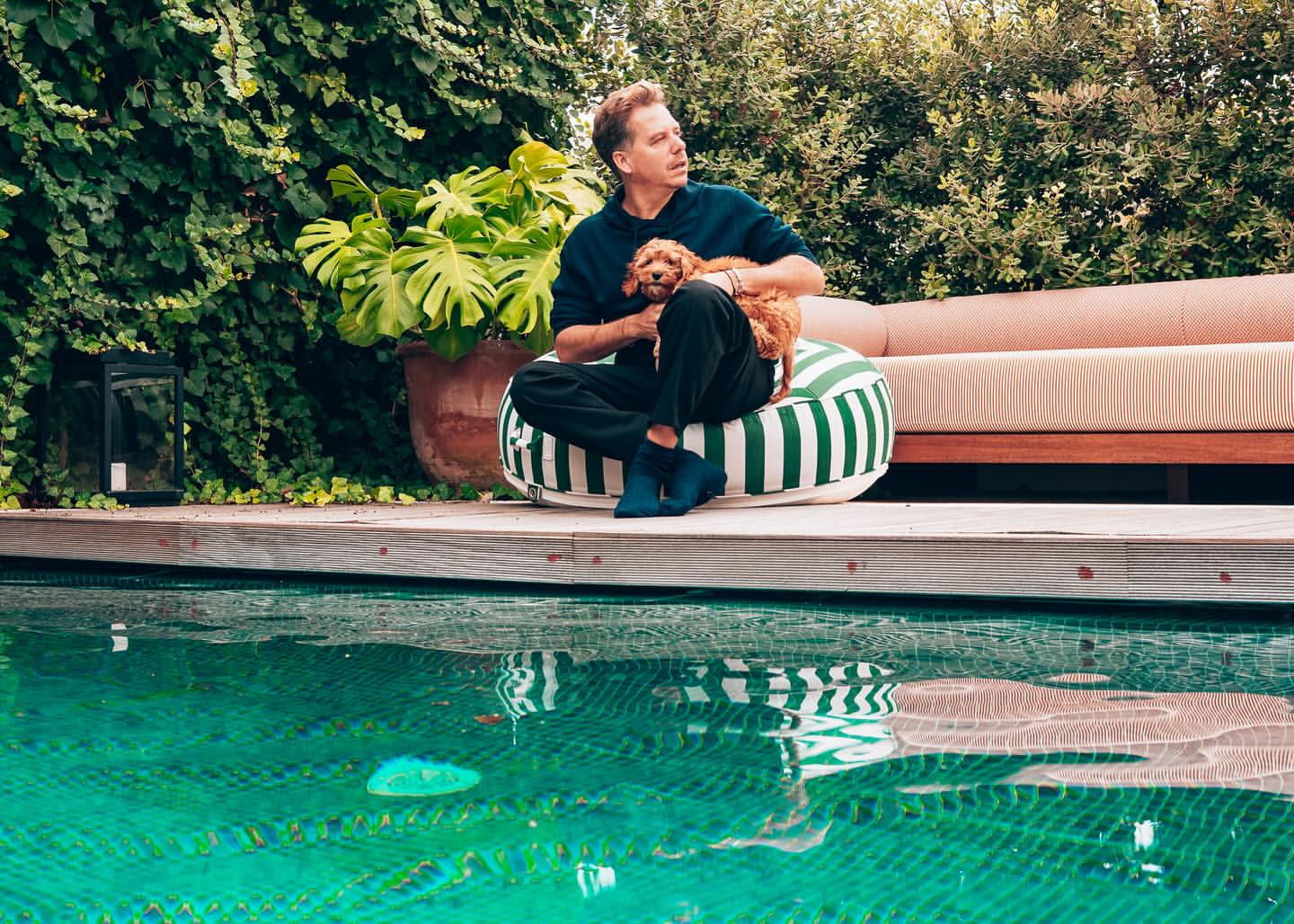Man and small dog rest near turquoise pool. Man wears navy, sits on striped cushion. Green foliage backdrop. Summer lifestyle scene.