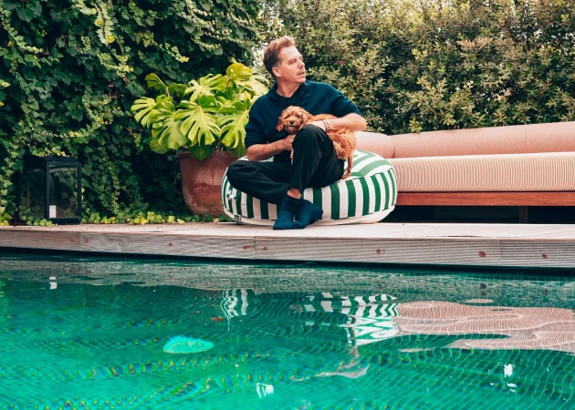 Man and small dog rest near turquoise pool. Man wears navy, sits on striped cushion. Green foliage backdrop. Summer lifestyle scene.